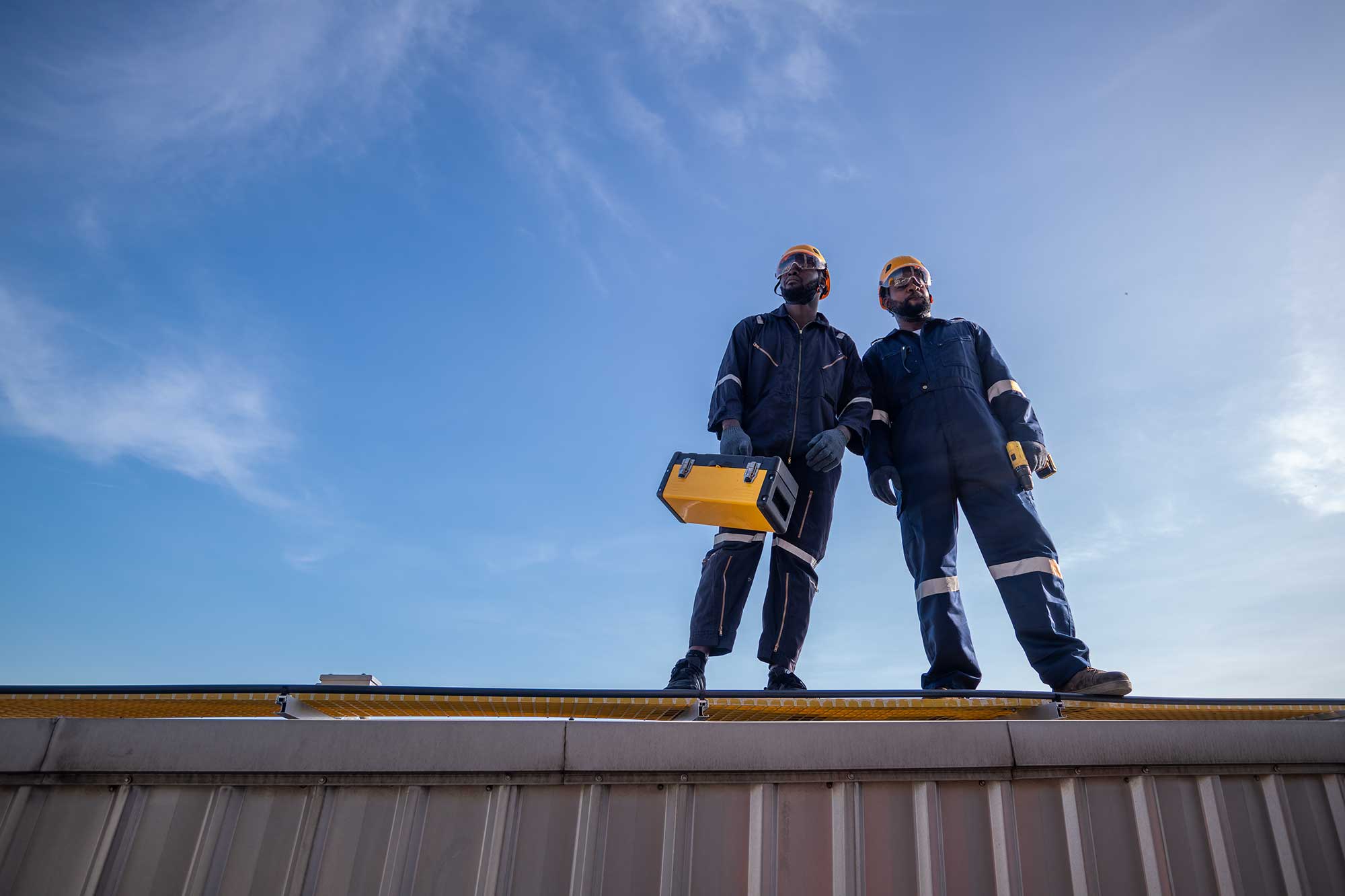 Two men in hard hats stand atop a building, looking out over the construction site and cityscape below.