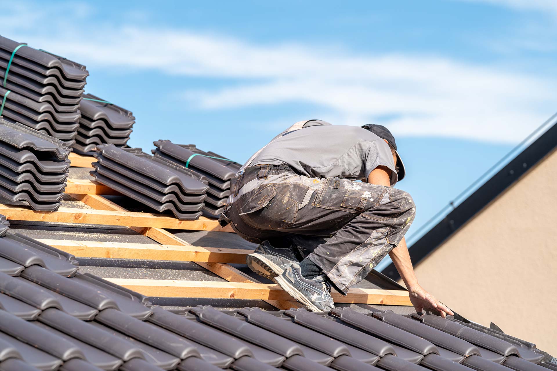 A man is working on a tiled roof, carefully adjusting tiles under a clear blue sky.