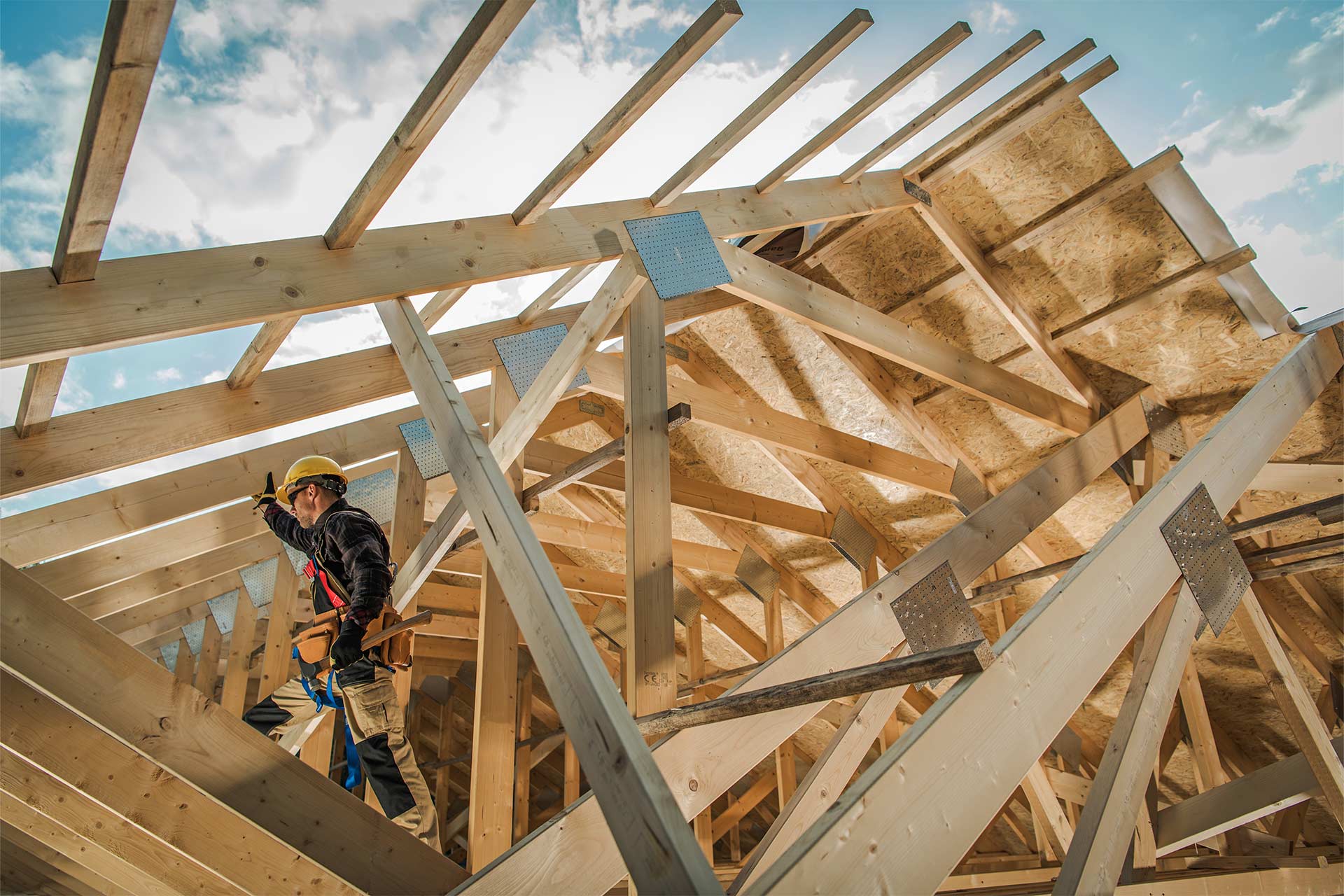 A construction worker gestures while navigating a wooden framework against a bright sky, showcasing intricate roof timber construction.