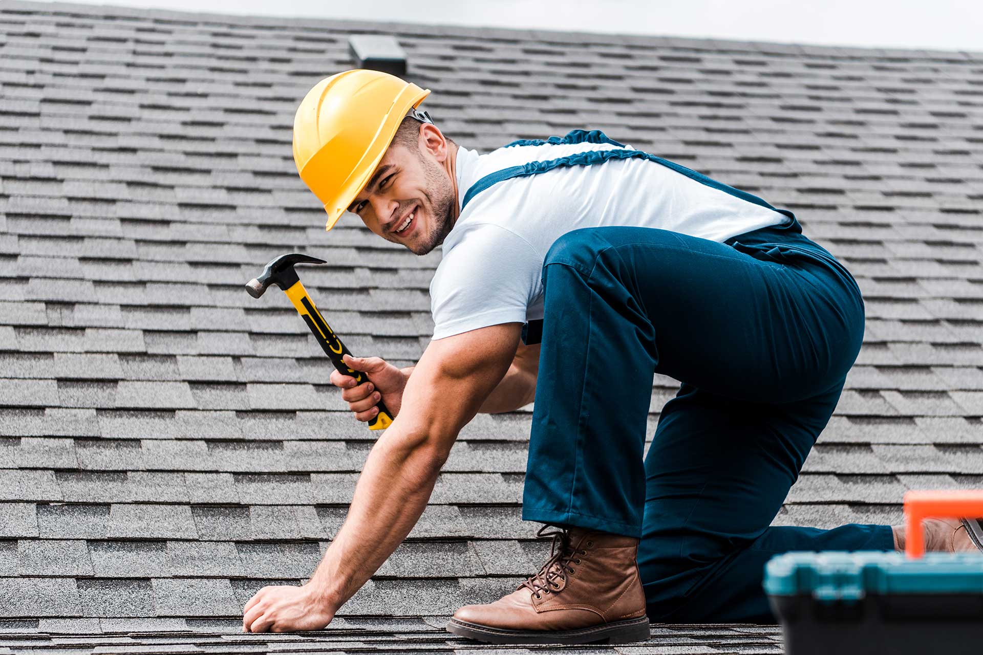 A construction worker in a yellow hard hat kneels on a shingled roof, holding a hammer, preparing to secure roofing materials.